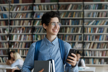 Happy dreamy young Jewish male student in eyewear holding books and cellphone in hands, looking in...