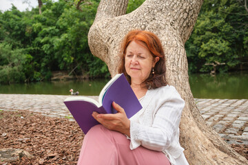 Mature hispanic woman reading a ebook in a park sitting against a tree.