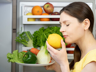 Obraz premium Young beautiful woman holds a lemon in her hand and sniffs it with her eyes closed, fruits and vegetables in the open fridge, woman takes fresh food from the refrigerator