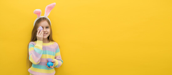 A little happy girl with long blonde hair in a headband with bunny ears holds colorful Easter eggs near her face and looks into the camera on a yellow background, an Easter card, an Easter banner, a