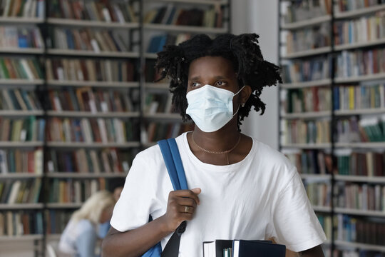 Head shot pensive young African American male student in protective facial mask, holding backpack and textbooks in hands, staying safe in public place library during pandemic situation, looking away.
