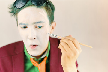 Mim is preparing for performance. Cosplayer paints his face with brush. Actor with white makeup and green hair in burgundy suit and yellow tie. Background with copy space for text.