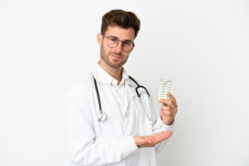 Young doctor caucasian man over isolated on white background wearing a doctor gown and holding pills