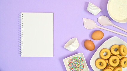 Creative copy space background with ingredients for baking cookies and open notebook for recipe on pastel purple background. Flat lay