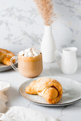 Croissant on a plate and delicious coffee with marshmallows in a cup on the table. Lifestyle homemade snack. Vertical view