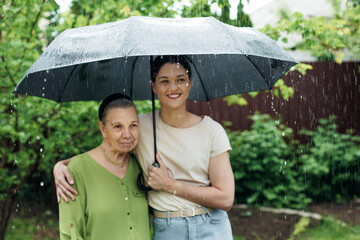 An old woman with her granddaughter stand outside in the rain with a black umbrella.