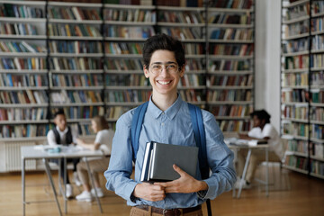 Portrait of smiling millennial Jewish male student in eyeglasses holing books in hands, posing in...