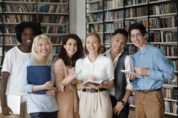 Portrait of smiling friendly smart millennial students with copybook in hands posing near bookshelves in modern library. Group of motivated young diverse friends ready for study, education concept.