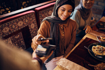 Happy Muslim woman paying contactless with mobile phone in restaurant. - obrazy, fototapety, plakaty