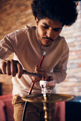 Young Muslim man adds hot coal cubes on narghile while smoking at home.
