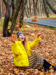 Laughing woman sitting in a park in a pile of autumn leaves throws up dry foliage