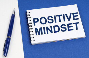 On a blue-white surface lies a pen and a notebook with the inscription - Positive Mindset