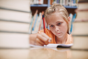 Hard at work. A cute young girl doing schoolwork while surrounded by books at the library.