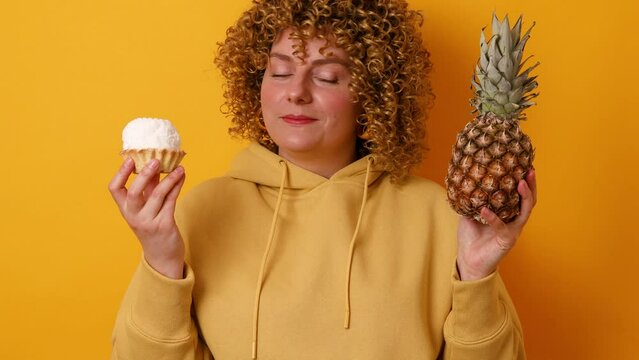 Young woman making choice between pineapple and tasty sweet cake isolated on yellow color background. Summer, diet and healthy lifestyle concept