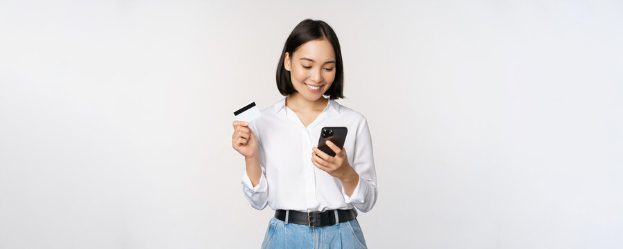 Online Shopping Concept. Korean Woman Holding Credit Card And Looking At Smartphone App, Buying, Order Delivery In Mobile Phone Application, Standing Over White Background