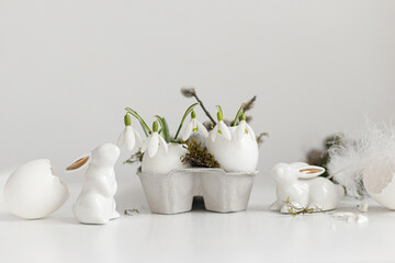 Easter rustic still life. Easter egg shells with blooming snowdrops, bunny figurines, feathers, nest on aged white wooden table. Simple stylish festive decoration on table. Happy Easter!