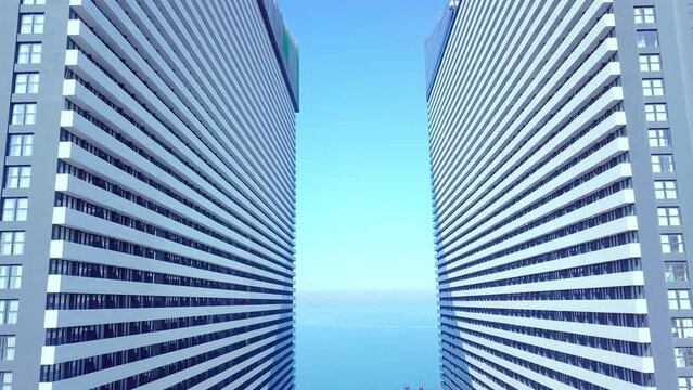 A close-up shot of the reflections of the sun, clouds and sky on the glass windows of a city skyscraper. The drone flies past through two buildings of a skyscraper on the background of sea or ocean.