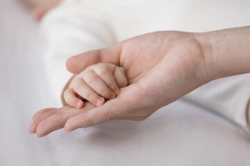 New mom touching tiny small palm of little baby. Mother holding cute hand of infant child wearing white garment clothes. Close up, cropped shot. Motherhood, childbirth concept