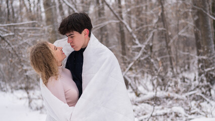Fototapeta premium A young couple walks in the park in winter. The guy and the girl are kissing wrapped in a white blanket outdoors.