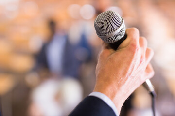 Closeup of male hand in formal suit holding microphone on blurred background of conference hall. Concept of speaking during business event