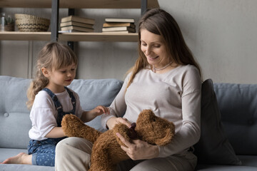 Positive engaged pregnant mom and adorable little daughter kid playing teddy bear toy on sofa, interacting in role games, enjoying home activity, playtime, entertainment, leisure. Motherhood, family