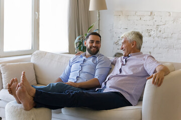 Grown up son and older father talking, share news enjoy warm communication pleasant conversation relaxing on cozy sofa in modern living room. Wellbeing multigenerational family understanding concept