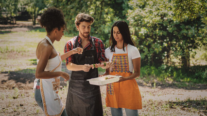 Group of three young people having fun on weekend cooking skewers on barbecue in the countryside