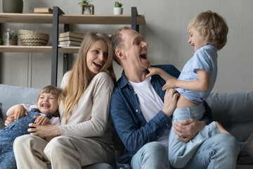 Joyful excited parents tickling funny little kids at home. Pregnant mom and happy dad playing active games with two siblings son and daughter, laughing, screaming, enjoying family leisure time - obrazy, fototapety, plakaty