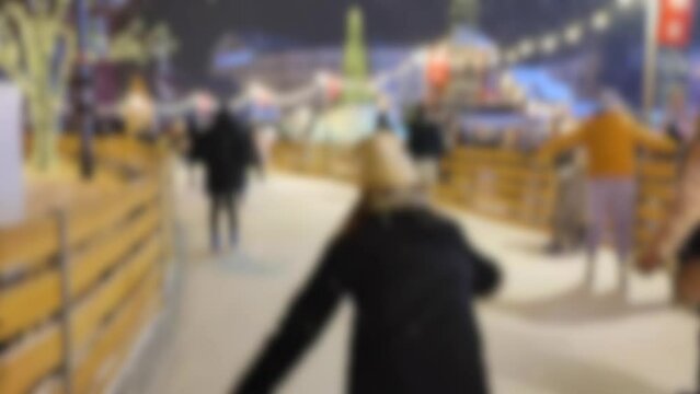 Blurred background. Many people skating on open-air ice skating rink. Snowfall on winter night. New Year Christmas. Street lights, luminous festive garland. Children family men women. City background