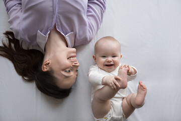 Happy cheerful mom and positive adorable few month baby resting together on bed mattress with white...