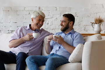 In living room happy senior dad and young adult son relax on couch drink coffee enjoy conversation, spend weekend together at home. Older younger gen relatives friendly relations, family ties concept