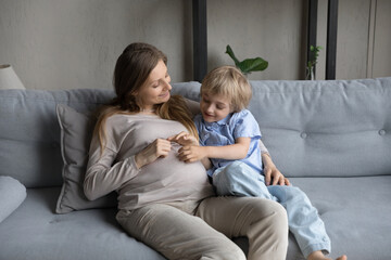Cheerful expecting mom and happy cute little son hugging, relaxing on couch together, touching...