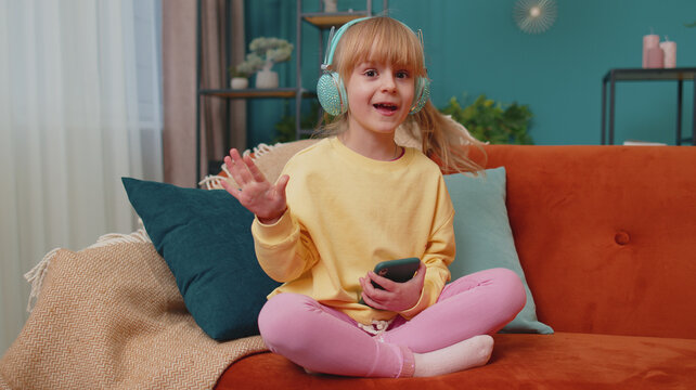Portrait of Caucasian funny little happy child girl sitting on sofa while listening music in headphones on smartphone player in living room. Toddler kid at home alone dancing, moving to rythm, singing