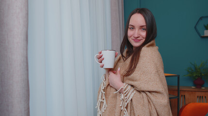 Portrait of smiling young brunette woman in warm white sweater and plaid drink hot beverage mug...