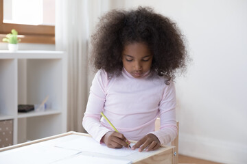 Concentrated cute little teenage curly African American kid girl drawing pictures with colorful pencils on paper alone at home, enjoying creative hobby activity, developing art skills sitting at table