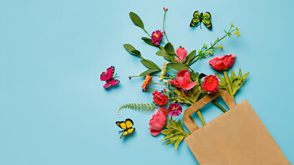Bouquet of various flowers in a shopping paper bag on pastel blue table background. Trendy flower delivery idea. Creative floral spring bloom concept. Still life natural visual trend. Flat lay.