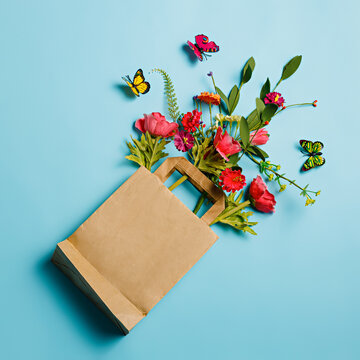 Bouquet of various flowers in a shopping paper bag on pastel blue table background. Trendy flower delivery idea. Creative floral spring bloom concept. Still life natural visual trend. Flat lay.