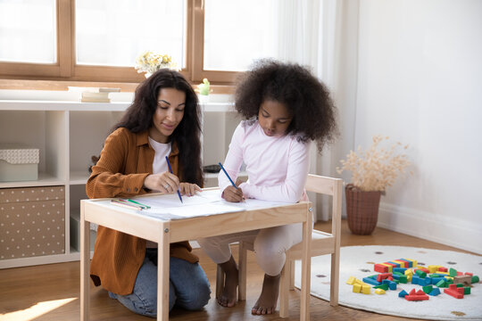Happy caring beautiful young African American mother or nanny teaching preteen child girl drawing with colorful pencils in paper album, enjoying creative early development activity together at home.