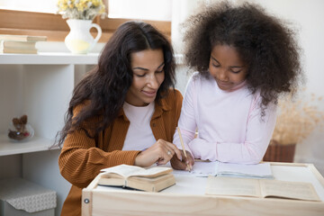 Caring young African American mother or nanny helping primary pupil cute little child daughter with school tasks, preparing homework together, learning writing on counting, sitting together at table.