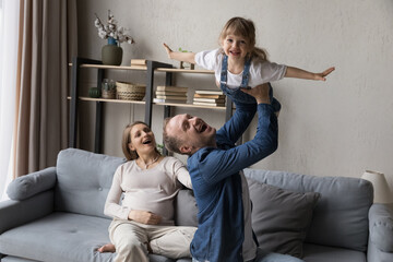 Excited expecting couple enjoying parenthood, playing with elder daughter kid at home. Happy dad lifting joyful girl up in air, child making flying airplane arms, laughing. Pregnancy, family concept