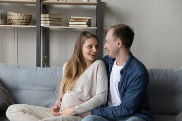 Happy young expecting couple enjoying leisure at home, sitting together on comfortable couch. Husband touching pregnant wife big belly with love, care, speaking, laughing. Family, pregnancy concept