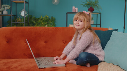 Little school kid girl use laptop computer sitting on sofa alone at home. Child learning reading online social media content, play education lessons game chatting with friends. Children tech addiction