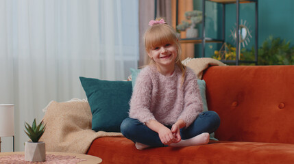 Portrait of carefree Caucasian little adorable happy child girl sitting on sofa in living room...