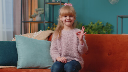 Ok. Portrait of Caucasian little happy child girl sitting on sofa at room looking approvingly at...