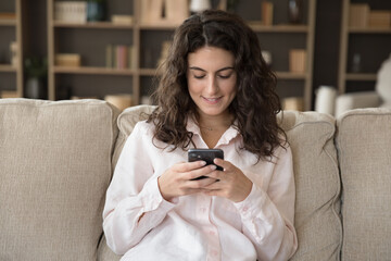 Relaxed attractive millennial curly Hispanic woman using cellphone applications, enjoying...
