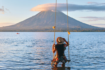 back view of a young girl sitting on a swing overlooking the volcano concenpcion on ometepe island,...