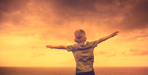 Happy little boy with arms outstretched facing a sunset background. Freedom and happiness concept 