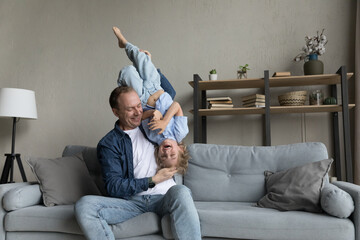 Happy cheerful dad holding excited funny preschool son kid in arms upside down, sitting on couch,...