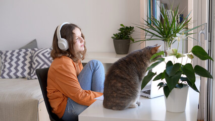 Relaxed teenage girl listening to music on headphones and looking out of the window with her cat