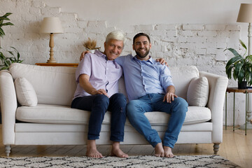Grey-haired elderly man his grown up young millennial 35s son sit on cozy couch in living room smiling looking at camera. Multi generational family portrait, ties and bonding, time and love concept
